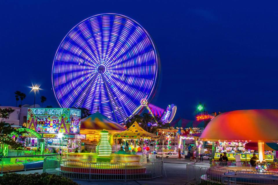 Wide night view of the park with the illuminated Ferris wheel above rides and booths.
