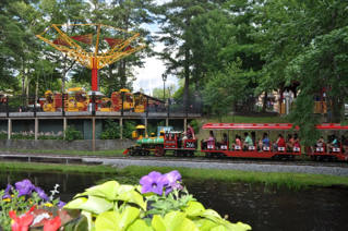 O Storytown Train passa junto ao lago florido, diante de uma atração aérea e edifícios entre as árvores.