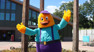 A mascote amarela Billy levanta os braços entre os pilares de madeira da entrada.